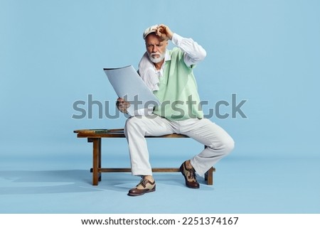 Portrait of handsome, serious, senior man in stylish outfit sitting on bench with sportive journal over blue background. Concept of leisure activity, hobby, lifestyle, fitness, emotions, retro style