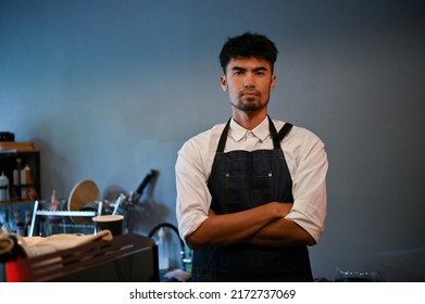 Portrait Of A Handsome, Smart, Millennial Asian Male Barista Or Coffee Shop Owner With An Apron Stands With Crossed Arms In The Coffee Shop.