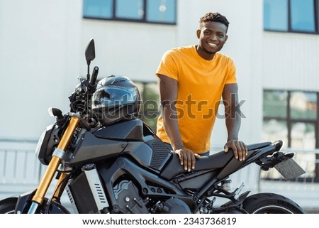 Portrait of handsome smiling african american man, biker looking at camera and posing near motorcycle at the street. Journey, freedom, transportation concept