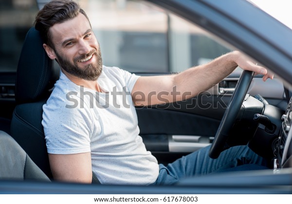 Portrait of a handsome smiling man\
dressed casual in white t-shirt sitting in the car in the\
city