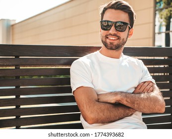 Portrait of handsome smiling stylish hipster lambersexual model.Man dressed in white T-shirt. Fashion male sitting on the bench in the street in sunglasses.Crossed arms - Powered by Shutterstock