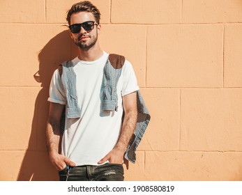 Portrait Of Handsome Smiling Stylish Hipster Lambersexual Model.Man Dressed In White T-shirt. Fashion Male Posing On The Street Background Near Wall In Sunglasses Outdoors