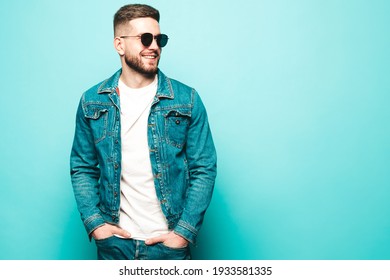 Portrait of handsome smiling stylish hipster lambersexual model.Man dressed in jacket and jeans. Fashion male posing near blue wall in studio in sunglasses