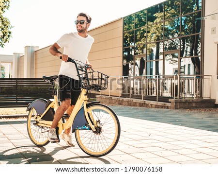 Portrait of handsome smiling stylish hipster lambersexual model.Man dressed in white T-shirt. Fashion male riding a bike on the street background in sunglasses