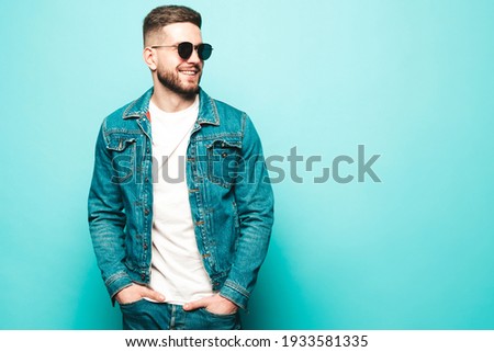 Portrait of handsome smiling stylish hipster lambersexual model.Man dressed in jacket and jeans. Fashion male posing near blue wall in studio in sunglasses