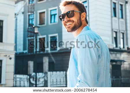 Portrait of handsome smiling stylish hipster lambersexual model.Modern man dressed in blue shirt. Fashion male posing near skyscraper on the street background in sunglasses. Outdoors at sunset 
