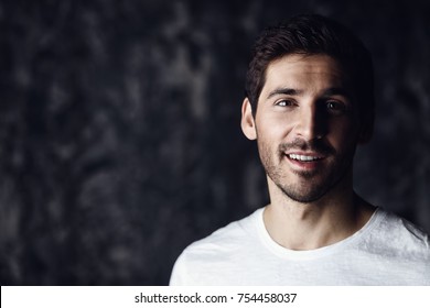 Portrait Of A Handsome Smiling Young Man Wearing White T-shirt. Studio Shot Over Dark Background. Men's Beauty And Health.