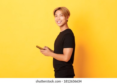 Portrait of handsome stylish asian guy in black outfit, using mobile phone and turning head at camera with satisfied smile, yellow background