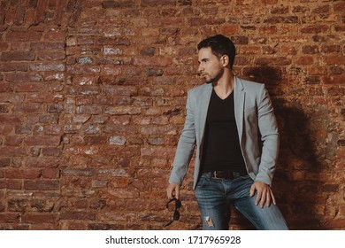 Retrato de un hombre bonito y elegante con chaqueta y jeans posando en el interior de un estudio de loft. Hombre de moda parado al lado de la pared de ladrillos mientras mira a un lado