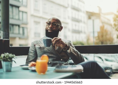 The portrait of a handsome stylish wealthy African guy with a beautiful black beard, in glasses, bald, in an elegant suit, sitting on a rainy morning in a street cafe and drinking delicious coffee