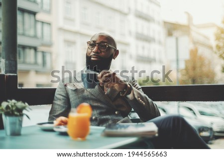 The portrait of a handsome stylish wealthy African guy with a beautiful black beard, in glasses, bald, in an elegant suit, sitting on a rainy morning in a street cafe and drinking delicious coffee