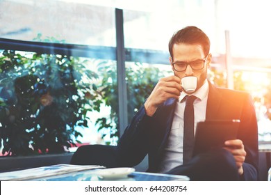 Portrait Of Handsome Successful Man Drink Coffee And Look To The Digital Tablet Screen Sitting In Coffee Shop, Business Man Having Breakfast Sitting On Beautiful Terrace With Plants