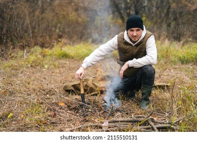 Portrait of handsome traveler male wearing warm clothes sitting near burning campfire and holding small shovel, looking at camera. Concept of bushcraft, camping and survival in nature. - Shutterstock ID 2115510131