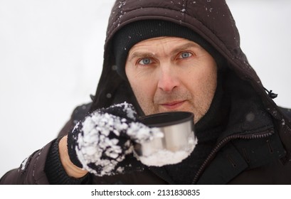 Portrait Of A Handsome Unshaven Man In Winter Clothes With A Hoodie Wearing Black Gloves Holding A Metal Mug. Portrait Of A Man 40-55 Years Old With A Mug Against A Background Of Snow.