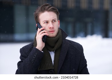 Portrait Of Handsome Upset Unhappy European Blonde Guy Young Nervous Man, Businessman Is Talking At Cell Mobile Phone Calling On Smartphone With Serious Face Outdoors At Cold Winter Snowy Day