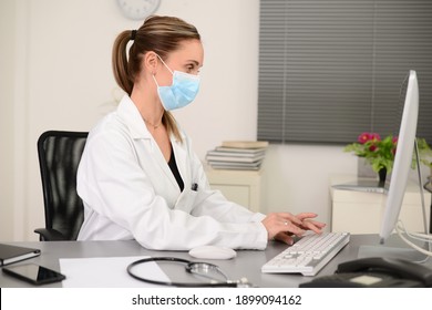 Portrait Of A Handsome Woman Female Doctor In Medical Practice Office Working On Computer Wearing A Surgical Mask