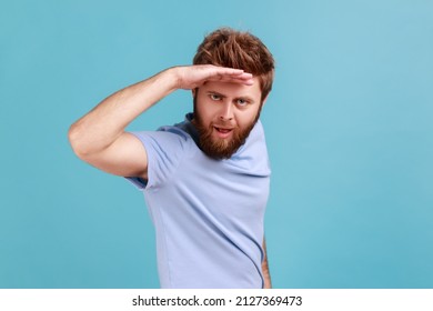 Portrait of handsome young adult bearded man looking far away at distance with hand over head, sees something pleasant with flirting expression. Indoor studio shot isolated on blue background.