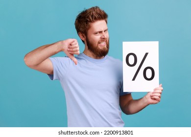 Portrait Of Handsome Young Adult Bearded Man Holding Paper With Percent Sign, Looking At Camera With Frowning Face And Showing Thumb Down. Indoor Studio Shot Isolated On Blue Background.