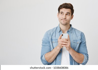 Portrait of handsome young adult with dreamy look, thinking while holding smartphone, isolated over white background. Son tries to made up message for his father, explaining why he took car - Powered by Shutterstock