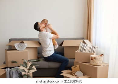 Portrait of handsome young adult man wearing white casual T-shirt and jeans, sitting on sofa with his baby daughter, father kissing his child, unpacking boxes after moving. - Powered by Shutterstock