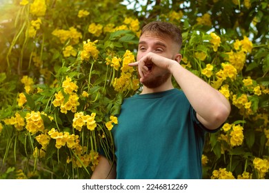 Portrait of handsome young allergic man is suffering from pollen allergy or cold on natural flowers, flowering tree background at spring or sunny summer day, sneezes, blowing his runny nose rubs eyes - Powered by Shutterstock