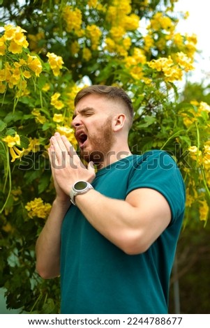 Portrait of handsome young allergic man is suffering from pollen allergy or cold on natural flowers, flowering tree background at spring or sunny summer day, sneezes, blowing his runny nose rubs eyes