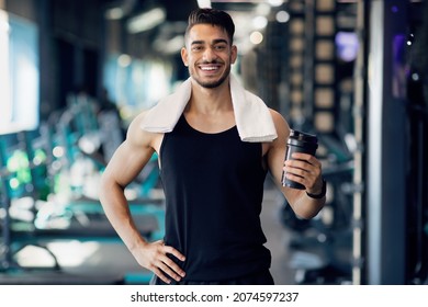 Portrait Of Handsome Young Arab Male Athlete Posing At Gym While Resting After Training, Middle Eastern Muscular Guy With Towel On Neck Holding Protein Supplement Shaker Bottle And Smiling At Camera - Powered by Shutterstock