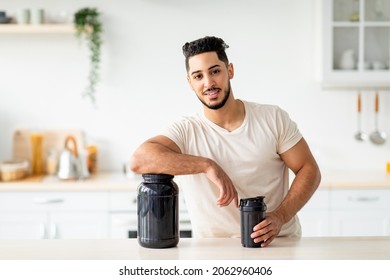 Portrait Of Handsome Young Arab Sportsman Posing With Protein Shake And Powder At Kitchen, Copy Space. Smiling Eastern Bodybuilder Using Sports Nutrition Product For Muscle Gain Or Weight Loss