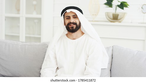 Portrait of handsome young Arabian man in kandura sitting on couch in living room and smiling to camera. Male Arab muslim in dishdasha on sofa at home. White interior Arabic millioner from Middle East