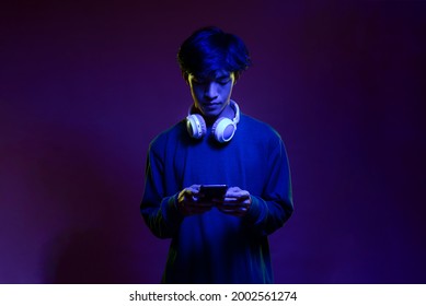 Portrait of handsome young asian man holding then looking at the mobile phone wearing headphone in neon light. Futuristic neon lighting. Trendy style - Powered by Shutterstock