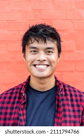 Portrait Of Handsome Young Asian Man Standing Outdoors Against A Red Wall Background In City And Looking At Camera - Happy Asian Guy Portrait - Generation Z People Concept