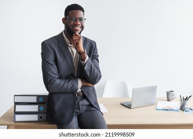 Portrait of handsome young black businessman in formal wear and glasses smiling at camera, sitting on work desk at modern office, empty space. Jobs and professions concept