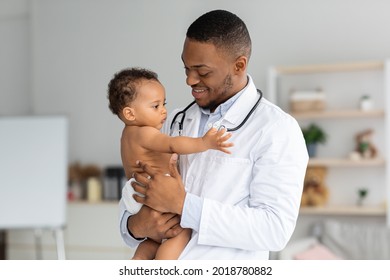 Portrait Of Handsome Young Black Pediatrician Holding Adorable Little Infant Boy In Hands, Cute Toddler Child Playing With Stethoscope On Doctor's Neck While Having Medical Check Up In Clinic