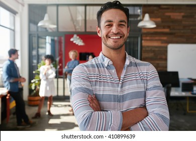 Portrait Of A Handsome Young Business Man Standing With Arms Crossed In An Office. Smart Caucasian Young Male Designer With His Colleagues In Background.