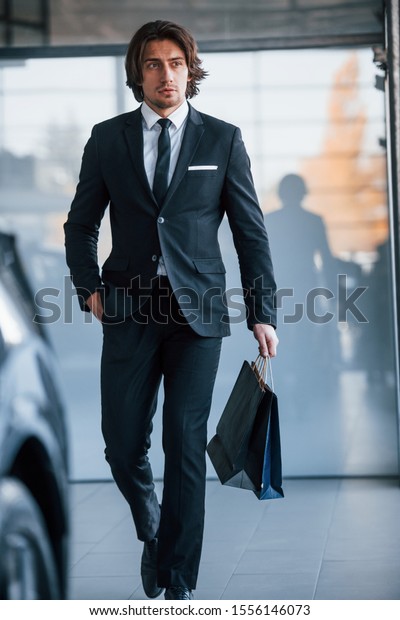 Portrait of handsome\
young businessman in black suit and tie and with shopping bag near\
the modern car.