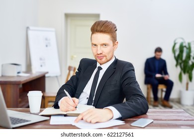 Portrait Of Handsome Young Businessman Looking At Camera With Smirk While Writing In Planner At Desk In Office, Employee In Background