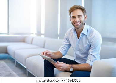 Portrait of a handsome young businessman sitting on a modern couch, holding a digital tablet, and smiling broadly at the camera