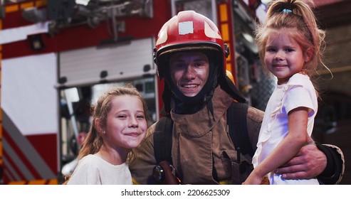 Portrait Of The Handsome Young Caucasian Fire Fighter Holding Two Small Saved Girls In Hands While They Looking At Camera And Happy Smiling Being Outdoors Next To Red Big Fire Truck Background.