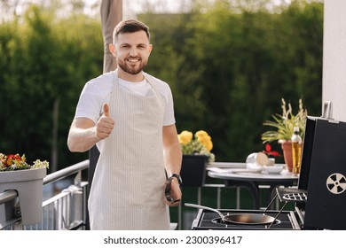 Portrait of handsome young chief in apron stand by the BBQ grill and smile. Male look into camera and showing thumbs up. - Powered by Shutterstock