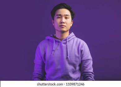 Portrait Of A Handsome Young Chinese College Student, Dressed In Casual Wear. Not Emotional Guy Looking At Camera With Serious Face, Isolated At Studio Against Puple Wall