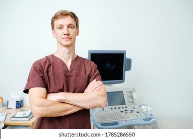 Portrait Of A Handsome Young Doctor Next To An Ultrasound Scanning Machine. He's Wearing Brown Shirt And Not The White Robe. Looking At The Camera.