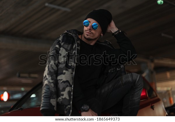 Portrait of a handsome\
young hipster guy with cool sunglasses in a fashionable winter\
military jacket with a black pullover and hat stands near a red car\
in the city
