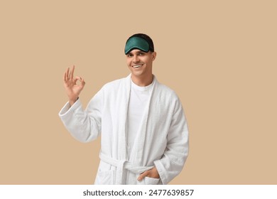 Portrait of handsome young man in bathrobe and sleeping mask showing OK on beige background