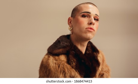 Portrait of a handsome young man in a fur coat. Gender fluid male with makeup and earring against beige background.