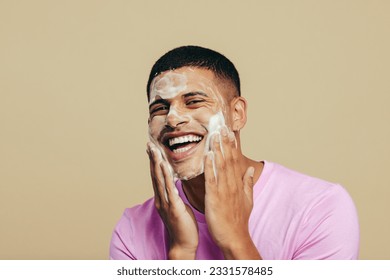 Portrait of a handsome young man indulging in his grooming and self-care routine as he applies face wash in a studio. Youthful man smiling happily while massaging his skin with a facial cleanser. - Powered by Shutterstock