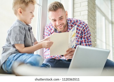 Portrait Of Handsome Young Man Reading Card For Fathers Day Sitting On Windowsill With His Son And Smiling Happily