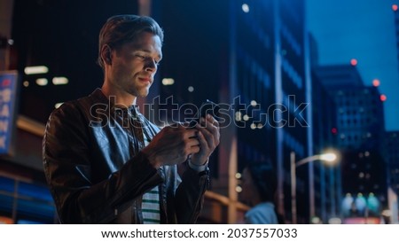 Portrait of Handsome Young Man Using Smartphone Standing in the Night City Street Full of Neon Lights. Smiling Stylish Blonde Male Using Mobile Phone for Social Media Posting.