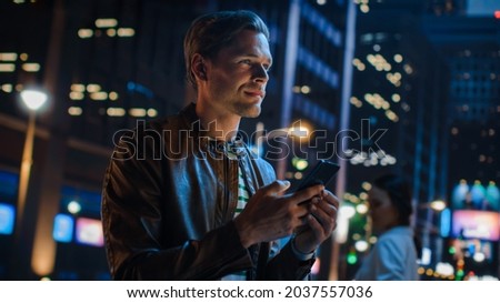 Portrait of Handsome Young Man Using Smartphone Standing in the Night City Street Full of Neon Lights. Smiling Stylish Blonde Male Using Mobile Phone for Social Media Posting. Dutch Angle Shot.