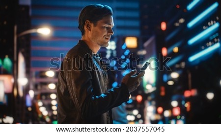 Portrait of Handsome Young Man Using Smartphone Standing in the Night City Street Full of Neon Lights. Smiling Stylish Blonde Male Using Mobile Phone for Social Media Posting.