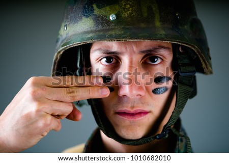 Portrait of handsome young soldier wearing a military uniform, painted his face with two fingers, in a gray background